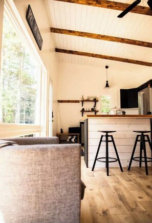 Una cocina con vigas de madera, una barra con taburetes, un refrigerador y una ventana con vistas a árboles.