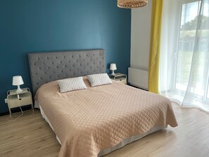Un dormitorio con cabecera capitonada, dos mesitas de noche con lámparas, una cama grande con cubierta acolchada y una ventana con cortinas de gasa.