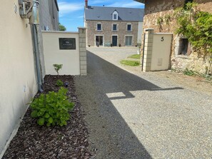 Un camino de grava conduce a un edificio de fachada de piedra, con una puerta marcada con el número 5 y un cartel que indica 'Gite au cœur du Marais'.