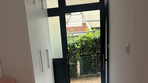Una habitación con sofá, armario y puerta que da a un espacio exterior con suelo de baldosas y vistas a un jardín.