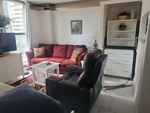 Ein Wohnzimmer mit einem roten Sofa, einem schwarzen Sessel, einem Fernseher, einem kleinen Tisch und einer Schrank mit Regalen.