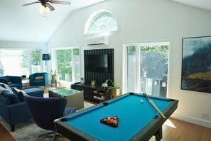 A living room with a pool table, blue sofas, a ceiling fan, and a large window.