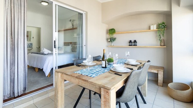 A dining area with a wooden table, chairs, and a bench. There is a sliding glass door leading to a bedroom.