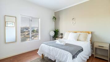 Une chambre à coucher avec un lit, une table de chevet, un attrape-rêves fixé au mur, une fenêtre avec des stores et un tableau encadré accroché au mur.