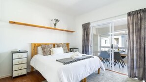 A bedroom with a bed, bedside tables, a wooden shelf, a window with curtains, and a view of a dining area.