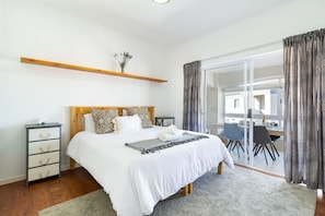 Quarto com cama, mesas de cabeceira, prateleira de madeira, janela com cortinas e vista para uma área de jantar.
