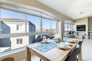 A dining area with a wooden table set for four, large windows, and a flat-screen TV mounted on the wall. - Yzer Drift Beach Escape 2 Bed 2 Bath (Yzerfontein)