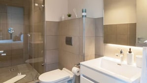 Un baño moderno con cabina de ducha de vidrio, inodoro blanco, lavabo con espejo y un pequeño cubo de basura.