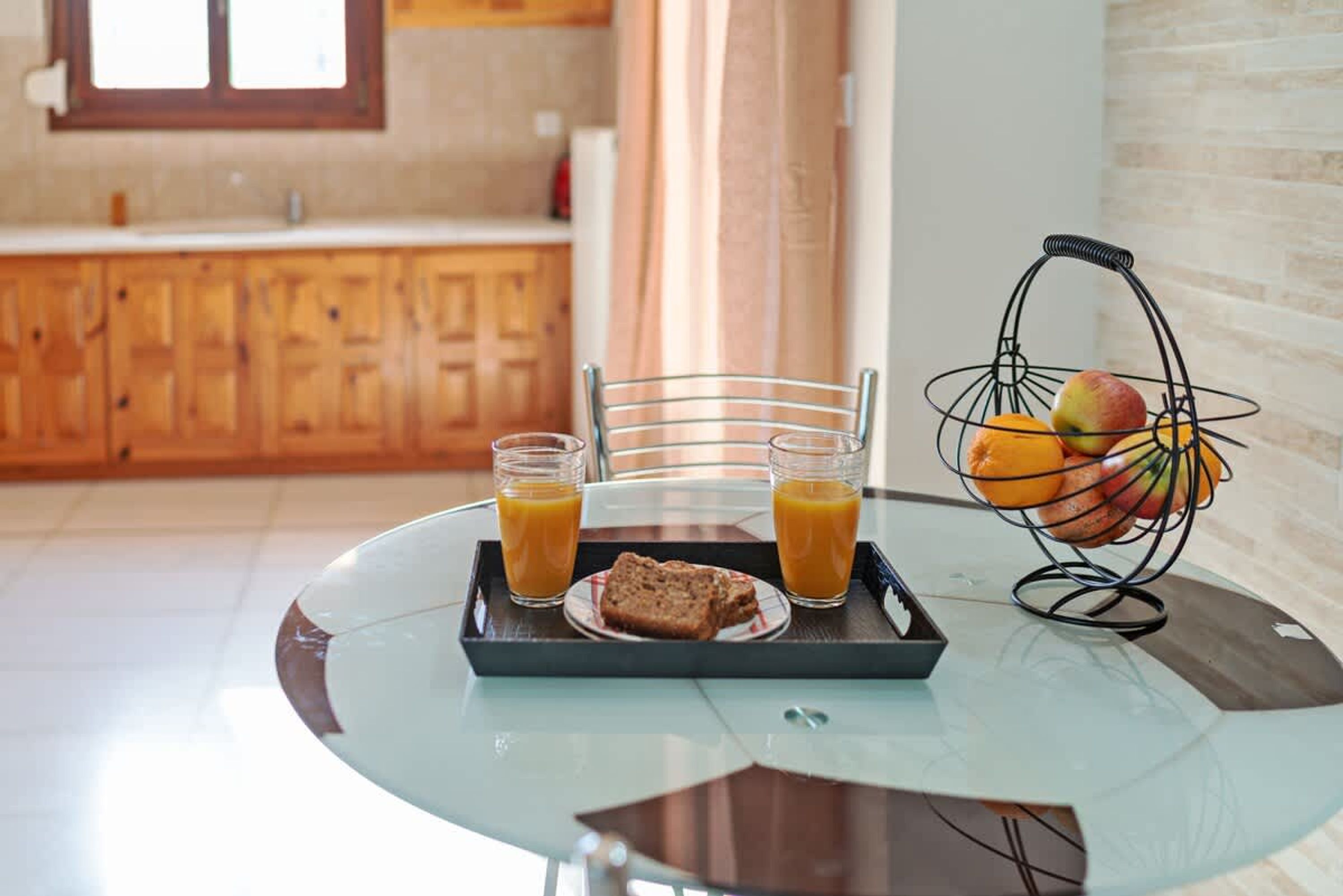 Bandeja de desayuno con vasos de jugo, tostadas y una cesta de frutas sobre una mesa de vidrio en una cocina con armarios de madera.
