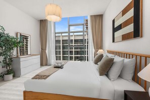 Ein modernes Hotelzimmer mit einem großen Bett, einem Nachttisch, einer Lampe, einer Zimmerpflanze und Blick auf die Stadt durch große Fenster.