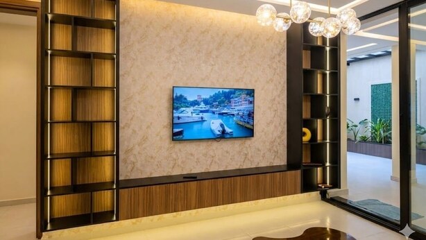 A modern living room with a flat-screen TV mounted on a textured wall, a built-in bookshelf, and a contemporary light fixture. - Spacious 3 Bedroom Apartment in Tangier - Near Stadium, Airport and Beaches (Tanger)