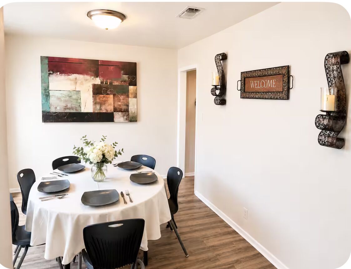 A dining area with a round table, black chairs, a vase with flowers, a 'WELCOME' sign, and abstract art on the wall.