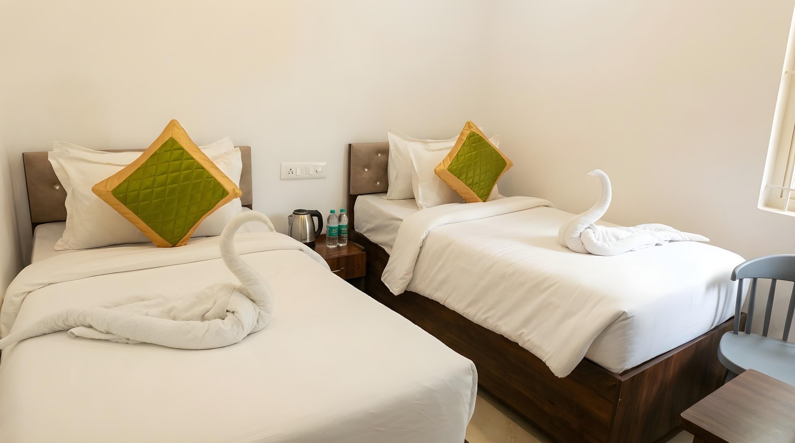 Two beds with white bedding and green quilted pillows, a nightstand with water bottles, and a chair.