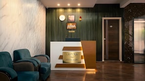 Um balcão de recepção com uma placa indicando 'OLIVRA HOTEL', um relógio, obras de arte emolduradas e uma porta com câmera de segurança.