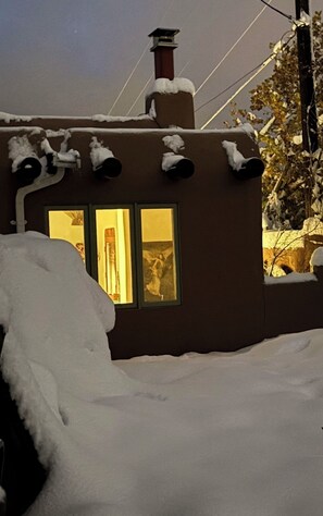 A house with a chimney, two lit windows, and snow on the roof and ground. - Casa de Monte Vista, balcony, patios, 3/3, 2 kit, 3 lvg areas, walk to Canyon Rd (Santa Fe)