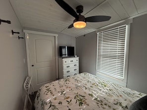 A bedroom with a bed, a dresser, a ceiling fan, a TV, and a window with blinds.