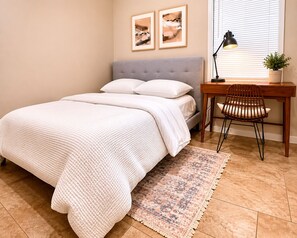 Une chambre à coucher avec un lit, un bureau avec une chaise, une lampe et deux tableaux encadrés accrochés au mur.
