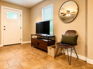 A room with a television on a wooden stand, a wicker chair with a cushion, a wicker basket, and a ceiling fan. - Pet-Friendly Central Austin Bungalow - Near Hospitals, Tech Corridor & Downtown (Austin)