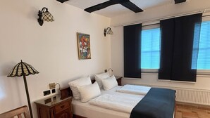 Een slaapkamer met een bed, nachtkastjes, een lamp, een schilderij, een raam met gordijnen en een betegelde vloer.