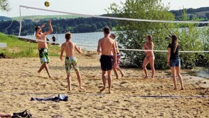 Partita di beach volley con giocatori in costume da bagno, una rete e un lago sullo sfondo.