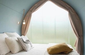 Ein Schlafzimmer mit einem Bett, Kissen, einem Fenster mit Vorhängen und einer Wandleuchte.