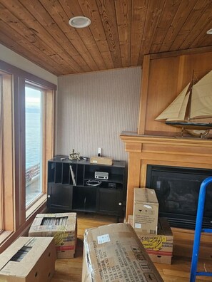 Huoneessa on puinen katto, takka, pienoismalli purjeveneestä ja laatikoita lattialla.