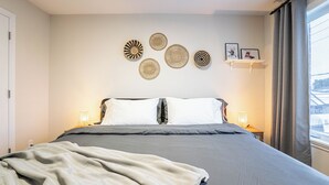 Uma cama bem arrumada com um cobertor cinza, uma parede com obras de arte em tear e uma janela com cortinas.