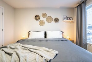 Ein ordentlich bezogenes Bett mit einer grauen Decke, eine Wand mit gewebten Kunstwerken und ein Fenster mit Vorhängen.