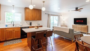 Een moderne keuken met houten kastjes, een centraal eiland, barstoelen, een eettafel, een bank en een televisie waarop 'NETFLIX' wordt weergegeven.