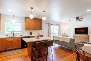 A modern kitchen with wooden cabinets, a central island, bar stools, a dining table, a sofa, and a TV displaying 'NETFLIX'. - 4BR | Sleeps 8, Fenced Yard, Games, King x 2 (Fargo)