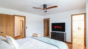 Een slaapkamer met een bed, een televisie waarop 'NETFLIX' staat, een plafondventilator en houten deuren.