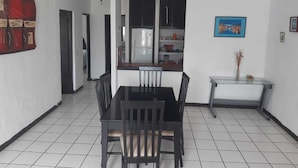A dining area with a black table and chairs, a ceiling fan, a refrigerator, and a kitchen area with cabinets and shelves. - Hotel Brisasol (Manzanillo)