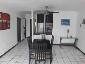 Un comedor con una mesa y sillas negras, ventilador de techo, refrigerador y una cocina con armarios y estantes.
