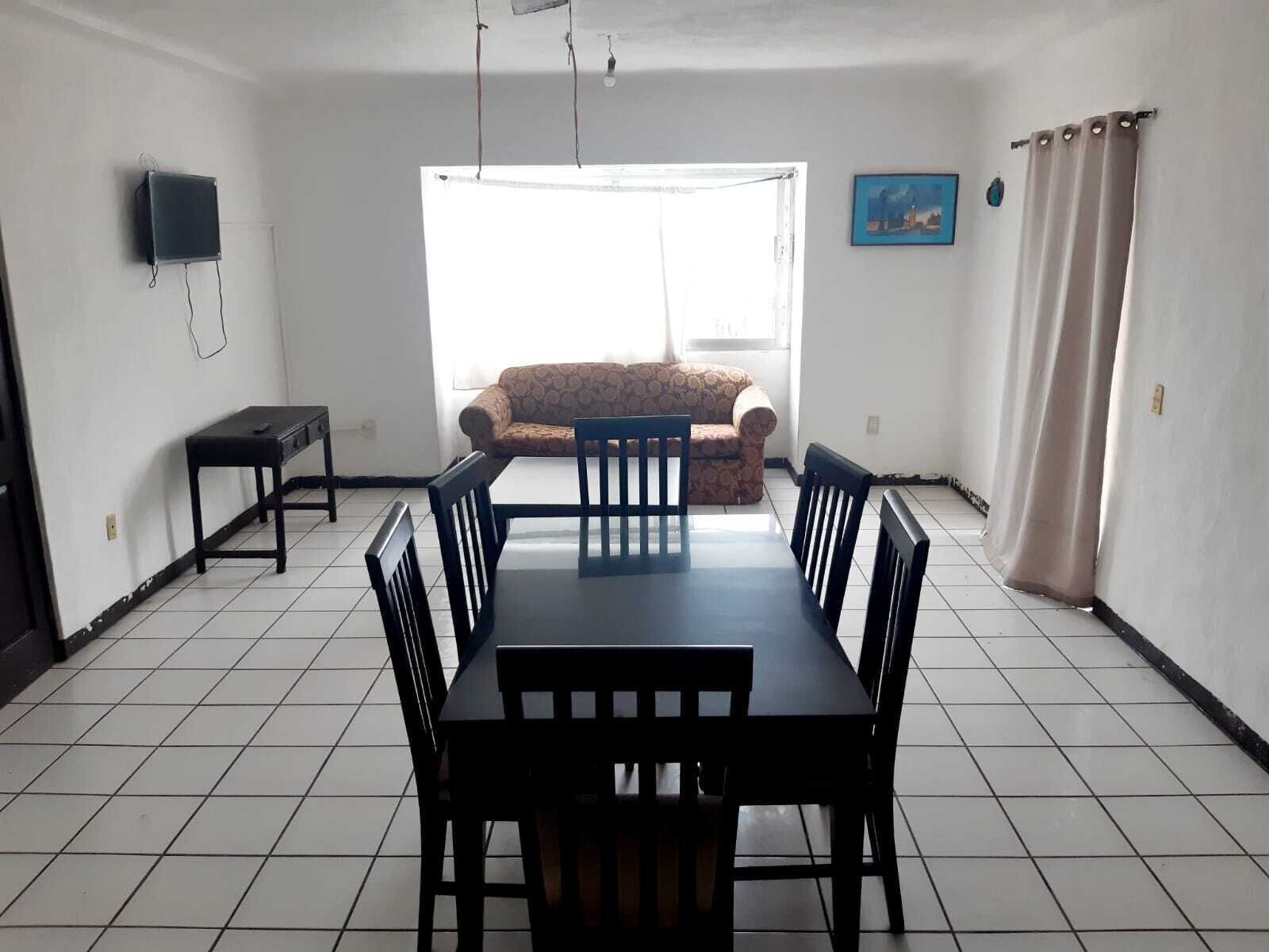 Un comedor con una mesa y sillas, un sofá, un televisor montado en la pared y una ventana con cortinas.