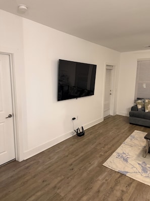 Ein Wohnzimmer mit einem an der Wand befestigten Flachbildfernseher, einem grauen Sofa und einem gemusterten Teppich auf Holzboden.