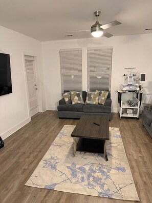 Ein Wohnzimmer mit niedrigem, dunkelholzigen Couchtisch, einem gemusterten Teppich, einem grauen Sofa mit Kissen, einem Deckenventilator und einer Nähmaschine auf einem Rollwagen.
