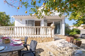 Ein weißes Gebäude mit einer Steinterrasse, Sitzgelegenheiten im Freien inklusive Stühle und einem Esstisch, sowie ein Balkon mit Geländer.
