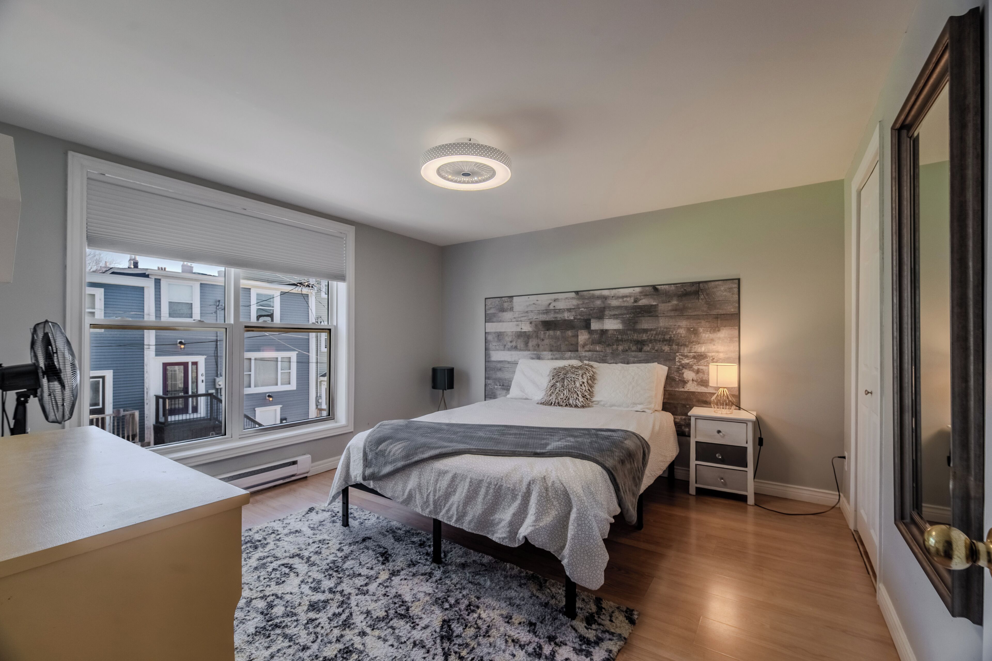 Een slaapkamer met een groot raam, een bed met een grijze deken, een nachtkastje met een lamp en een kunstwerk aan de muur.