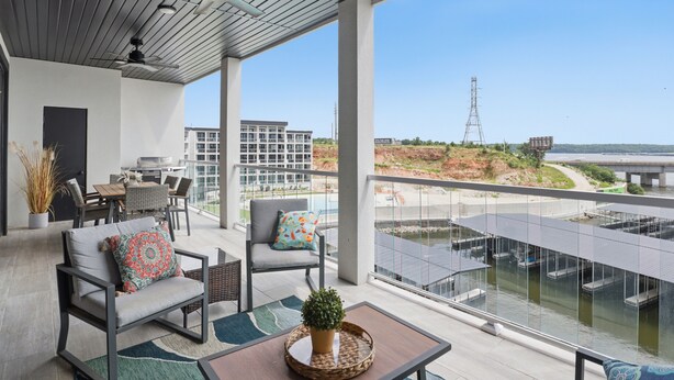 Un balcón moderno al aire libre con asientos, una zona de comedor y vistas a un cuerpo de agua y edificios distantes.