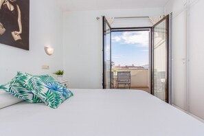 Een slaapkamer met een bed, een wandlamp, een kamerplant en uitzicht door een open schuifdeur op een buitenruimte met een tafel en stoelen.