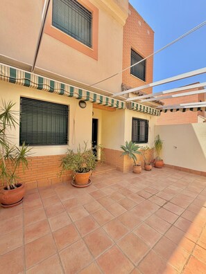 A courtyard with tiled flooring, potted plants, a covered area with a striped awning, and a brick building with windows. - TERRASOL HOUSE CERRO DEL ÁGUILA (Torre del Mar)