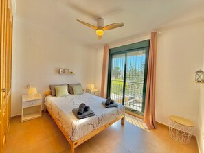 A bedroom with a bed, bedside tables, a ceiling fan, a window with curtains, and a view of the outside.
