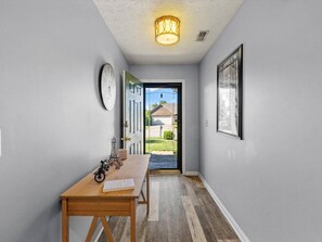A hallway with a wooden table, a clock, a door leading outside, and a framed picture on the wall.