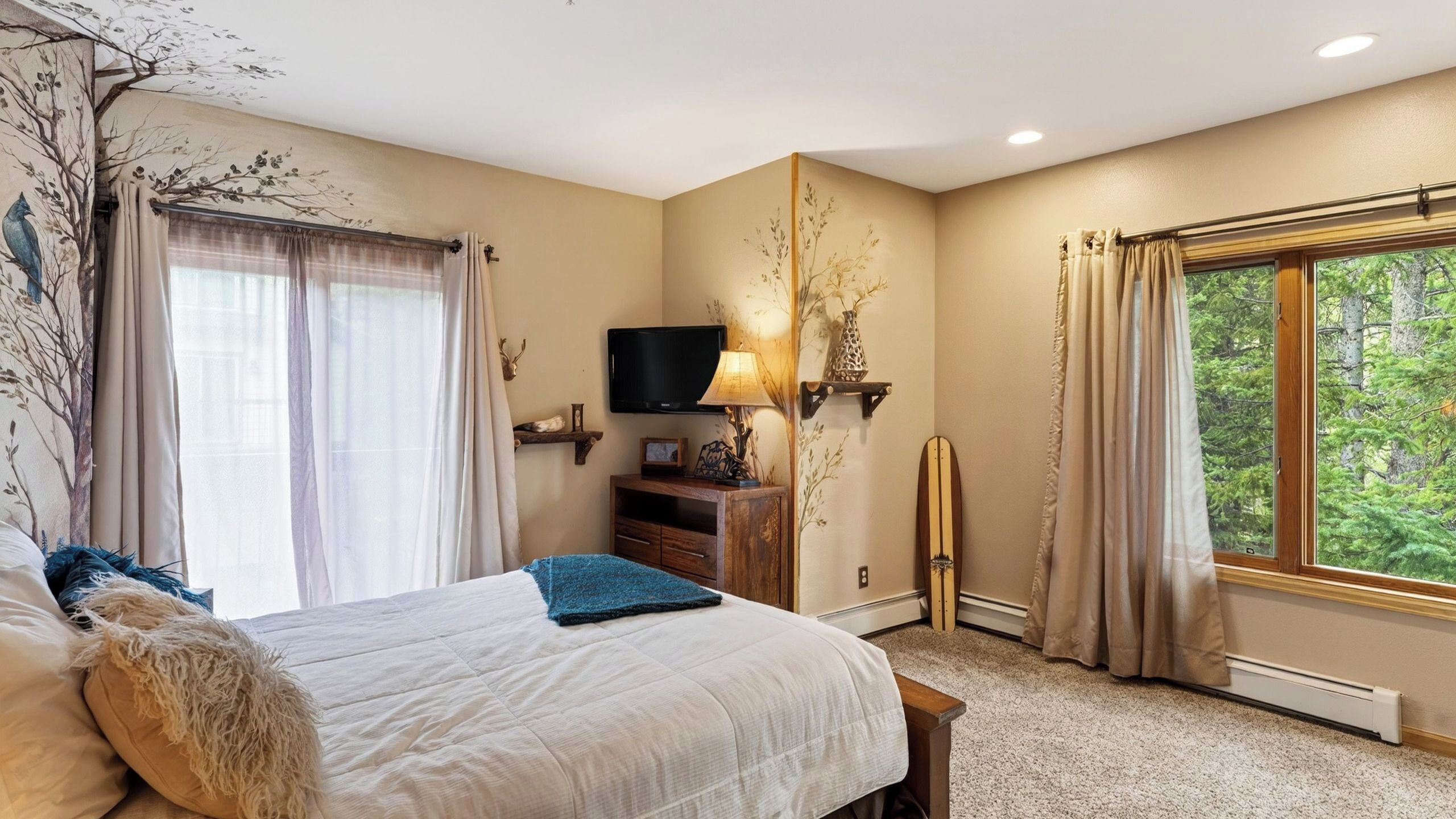 A bedroom with a bed, a TV, a lamp, a dresser, and a window with a forest view.