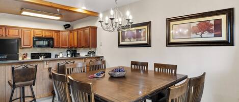 A dining area with a wooden table and chairs, a chandelier, framed pictures on the wall, and kitchen appliances in the background.