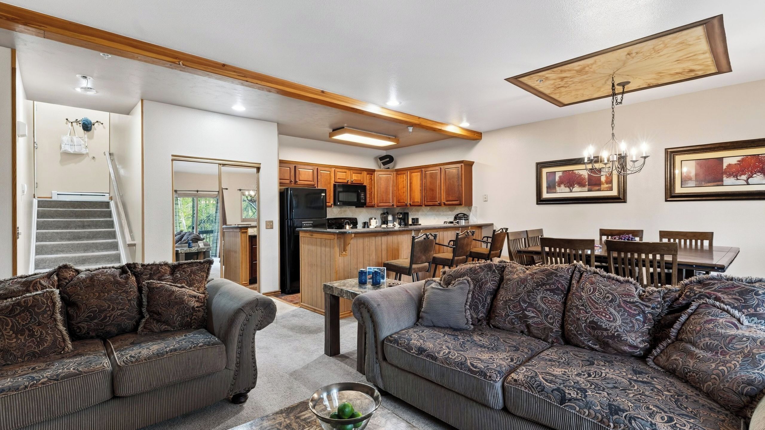 A modern living room with a sofa set, a dining table with chairs, a kitchen area with wooden cabinets, and a staircase leading to another level.