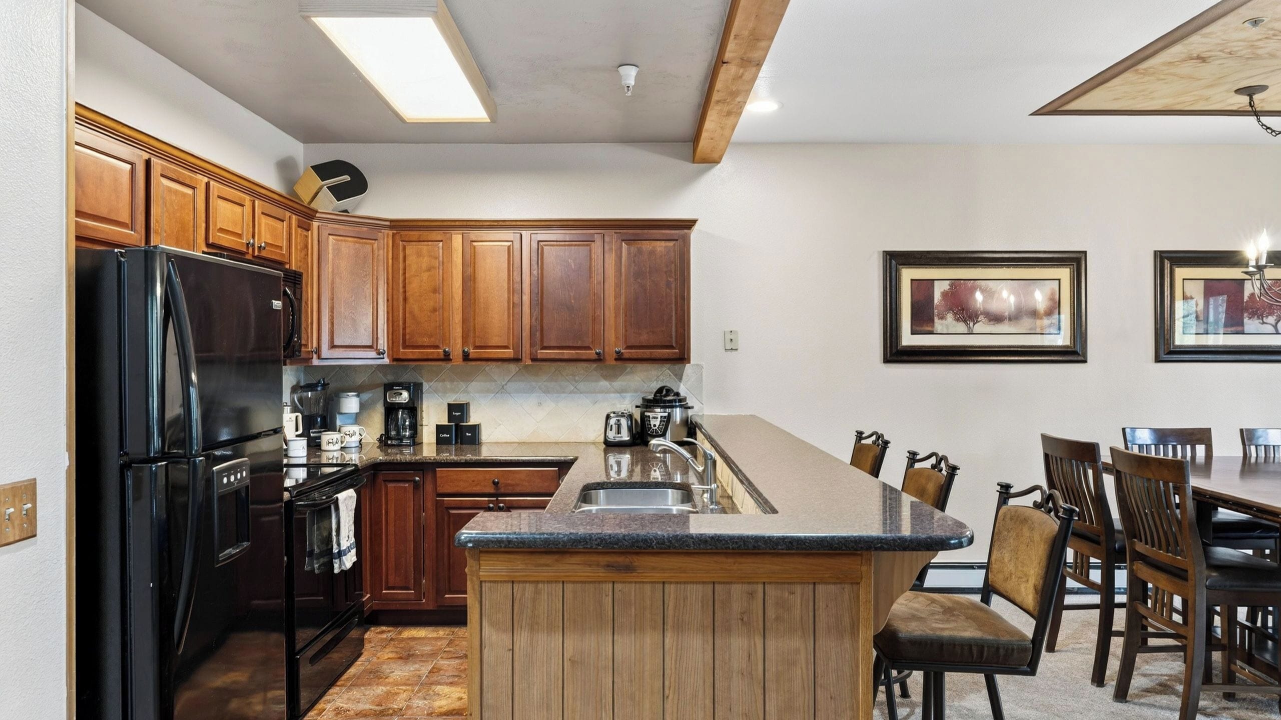 Una cocina con armarios de madera, un refrigerador negro, una cafetera y un comedor con sillas y una mesa.