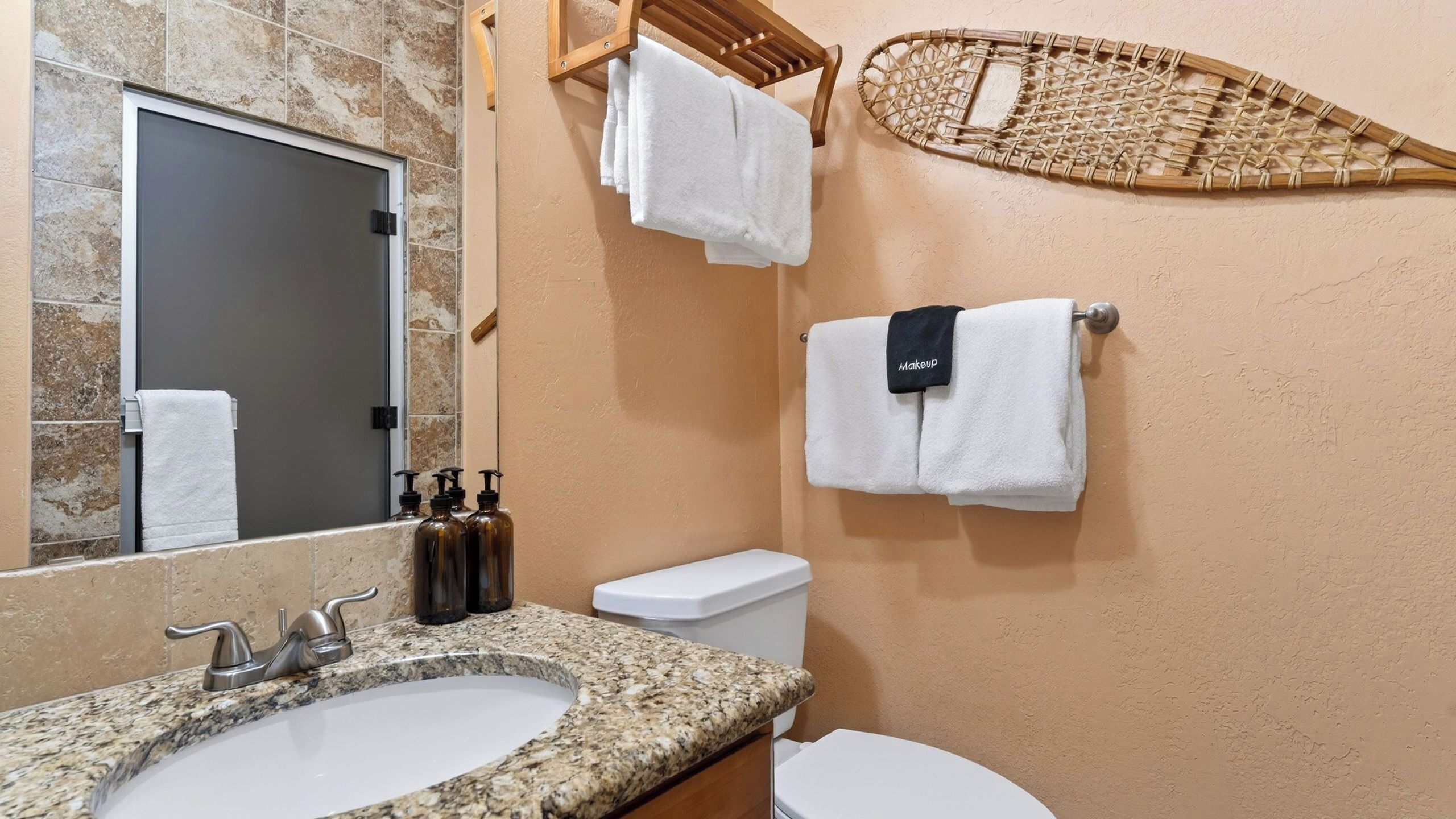 Une salle de bain avec un lavabo doté d’un plan de travail en granit, des toilettes blanches, un miroir et un porte-serviettes avec plusieurs serviettes.
