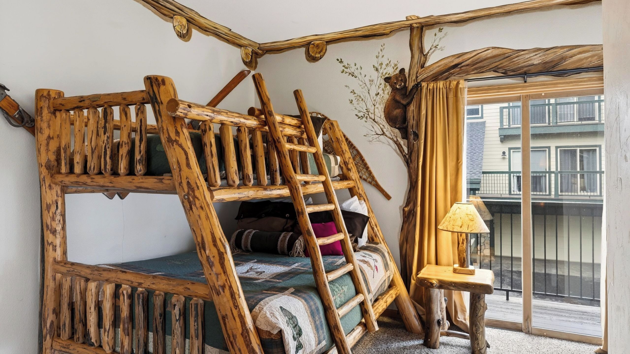 A rustic wooden bunk bed with a ladder, a small bedside table with a lamp, and a view of a balcony with a railing.