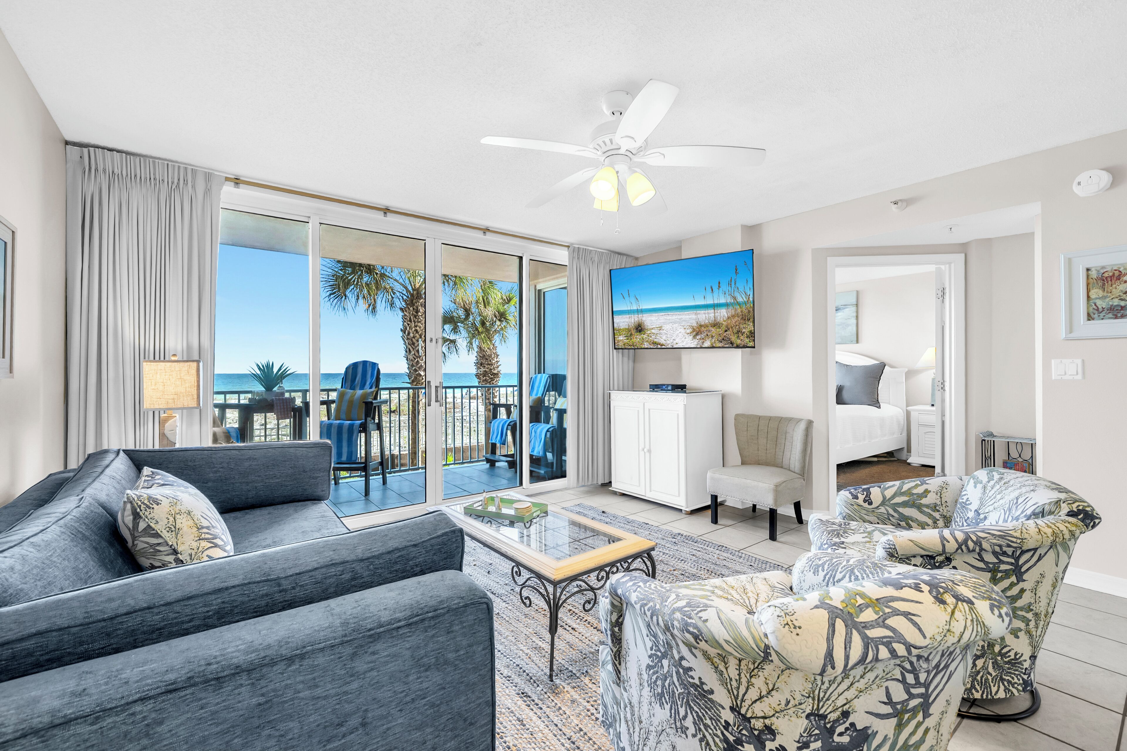 A modern living room with a sofa, armchair, and a coffee table. There is a large window with a view of palm trees and the ocean.